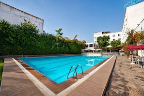 A swimming pool with a wooden deck, surrounded by greenery and a white building with a sign. - Hotel Mamora (Kenitra)