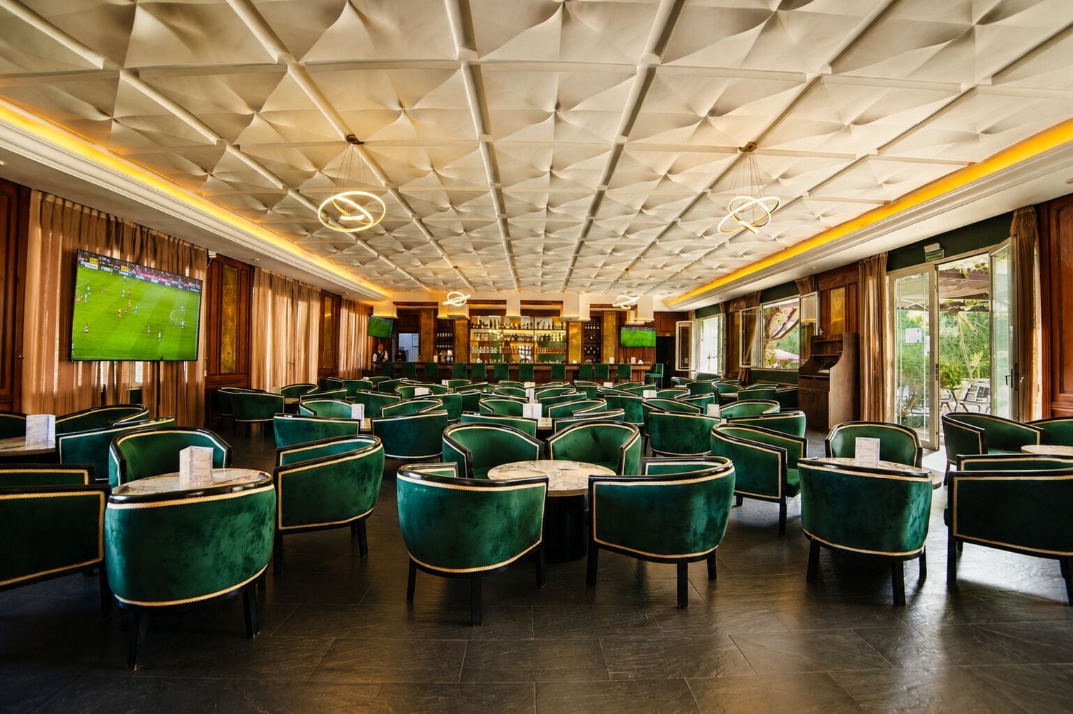 Un hall d’hôtel spacieux avec un plafond haut, des fauteuils à revêtement vert, un coin bar et un écran de télévision diffusant un événement sportif.