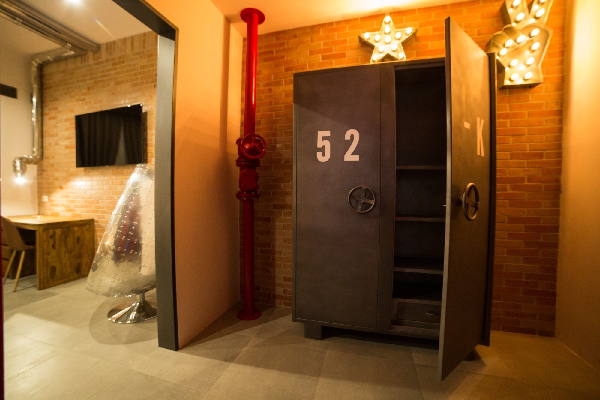 A hotel room with a brick wall, a number 52 safe, and a red pipe.