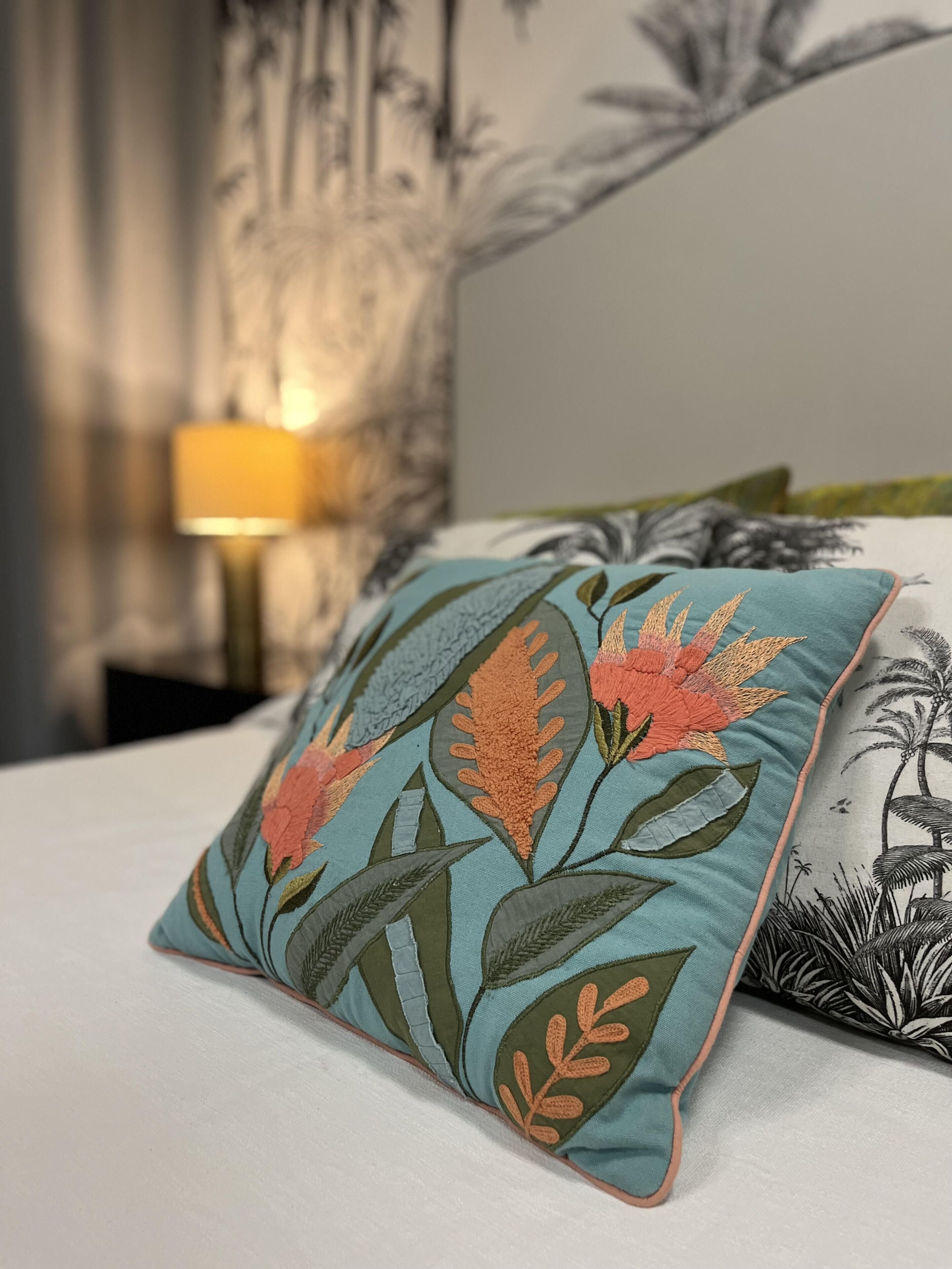 A bed with a teal pillow featuring floral embroidery, a white pillow with a palm tree design, and a lamp in the background.