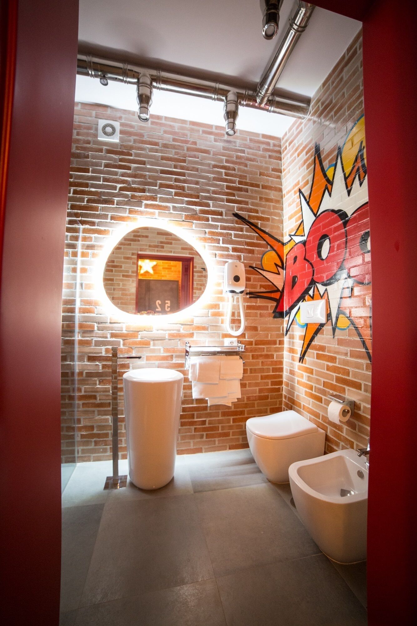 A bathroom with a brick wall, a round mirror, a toilet, a bidet, and a sink.