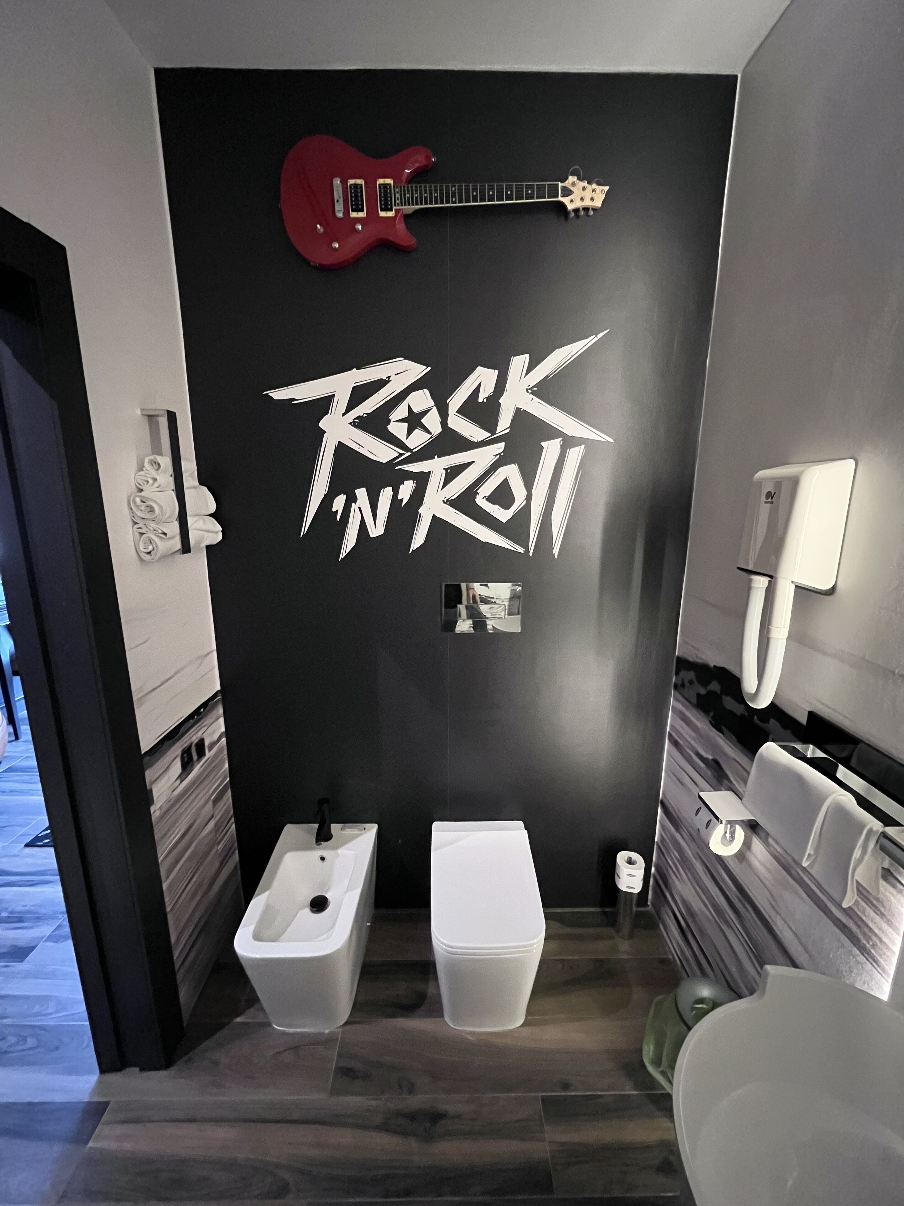 A bathroom with a black wall featuring the text "Rock 'n' Roll" and a red electric guitar. There are two sinks and a toilet.