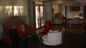 A spacious living area with a red sofa, a white armchair, and a black armchair. There is a glass coffee table and a small white table. The room has a large window with sheer curtains and a view of the outside. - Ozcelik Hotel (Salihli)