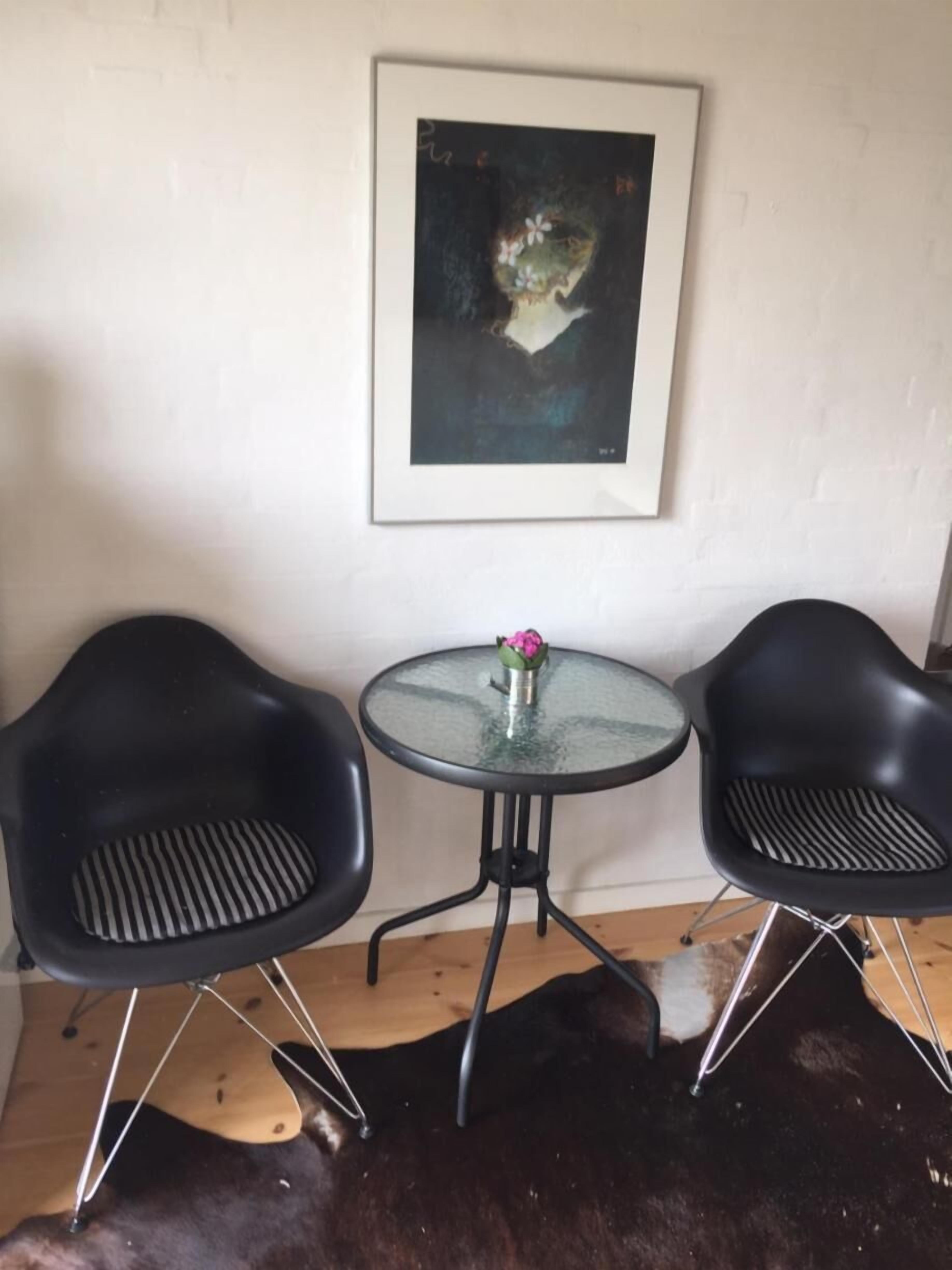 A room with a round glass table, two black chairs, a framed painting on the wall, and a dark brown rug.