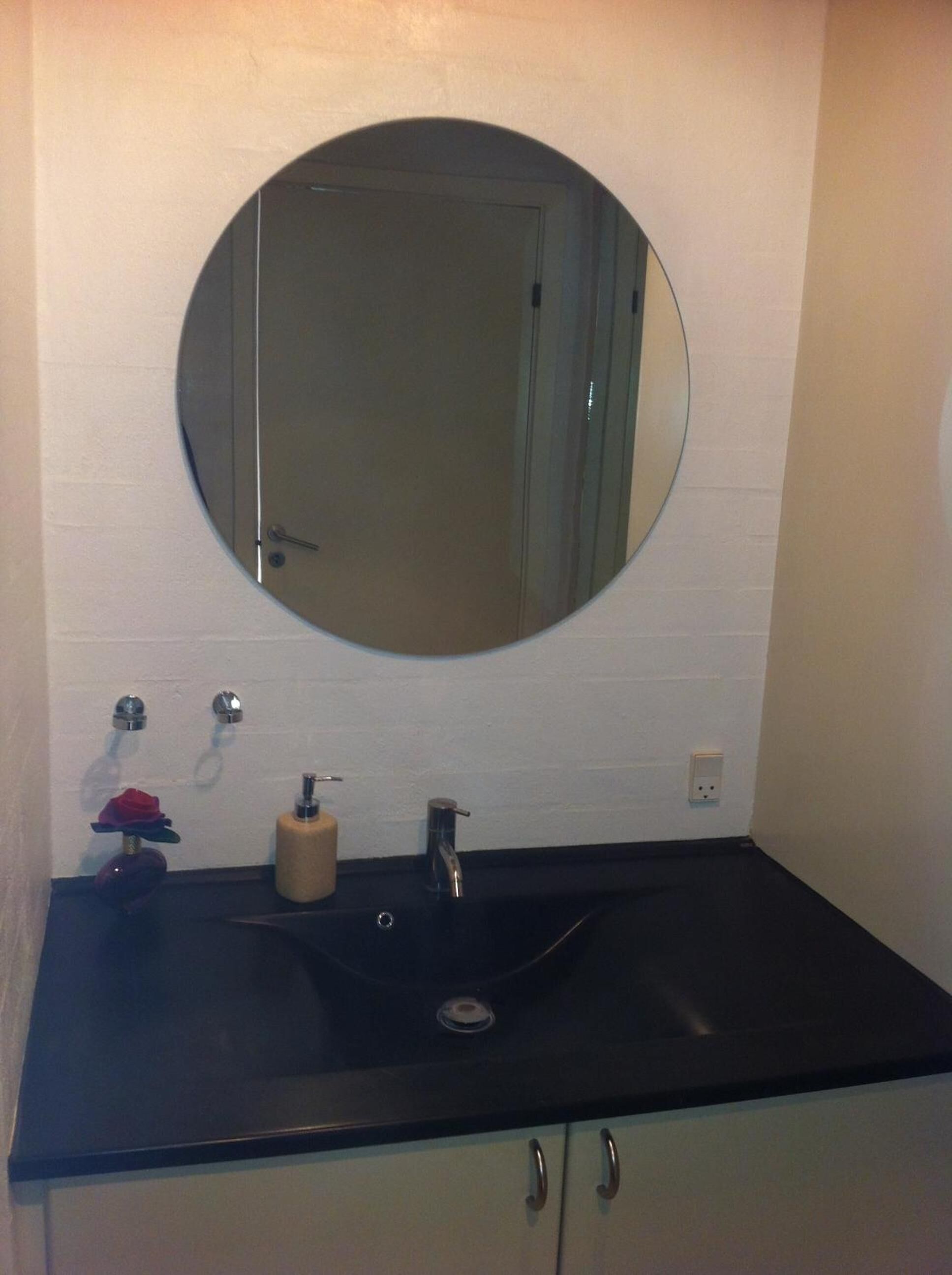 Un baño con un espejo redondo, un lavabo negro y una pared de azulejos blancos.