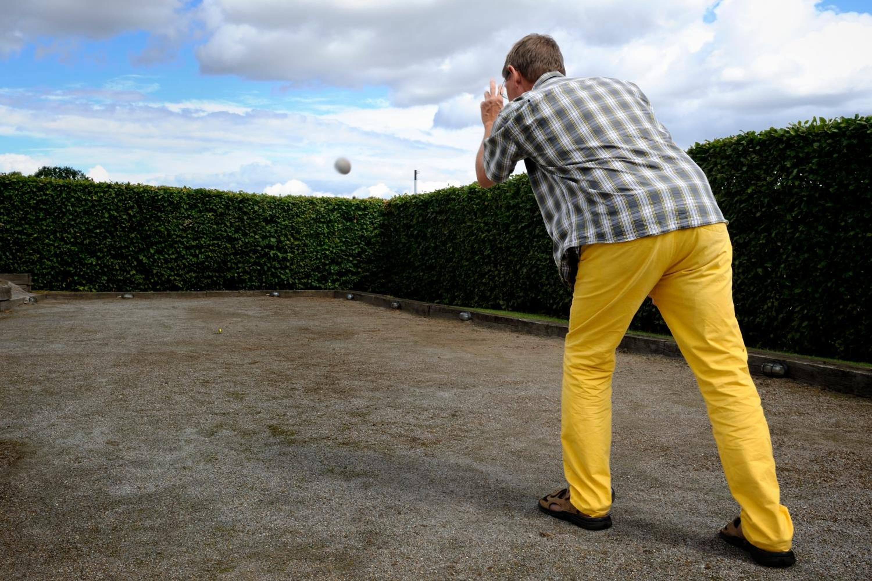 Un hombre con camisa de cuadros y pantalones amarillos lanza una pelota en un espacio al aire libre con seto y cielo despejado.