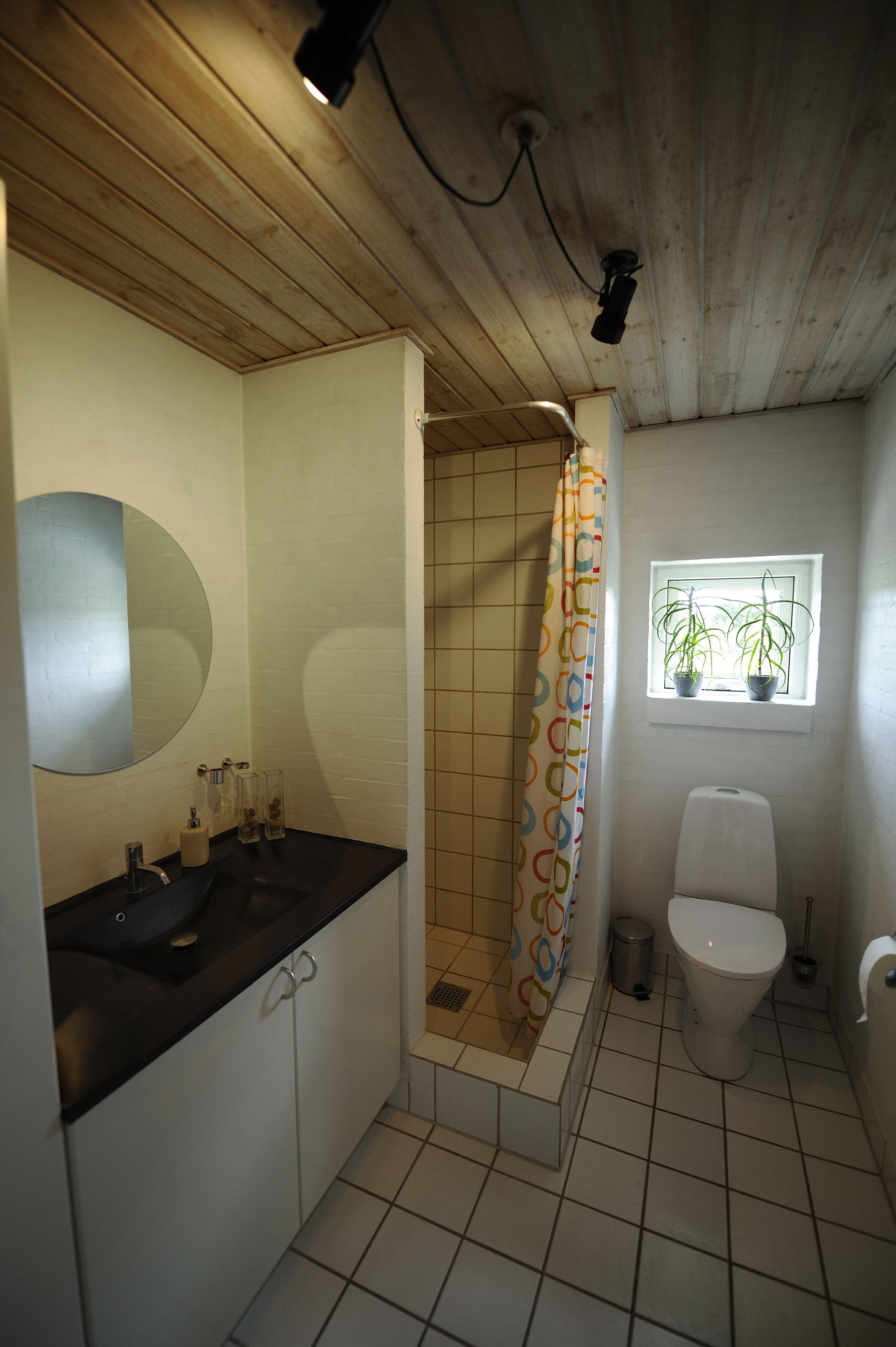 Une salle de bain avec un plafond en bois, un lavabo noir, un miroir, une douche avec un rideau coloré, des toilettes et une fenêtre ornée de plantes.