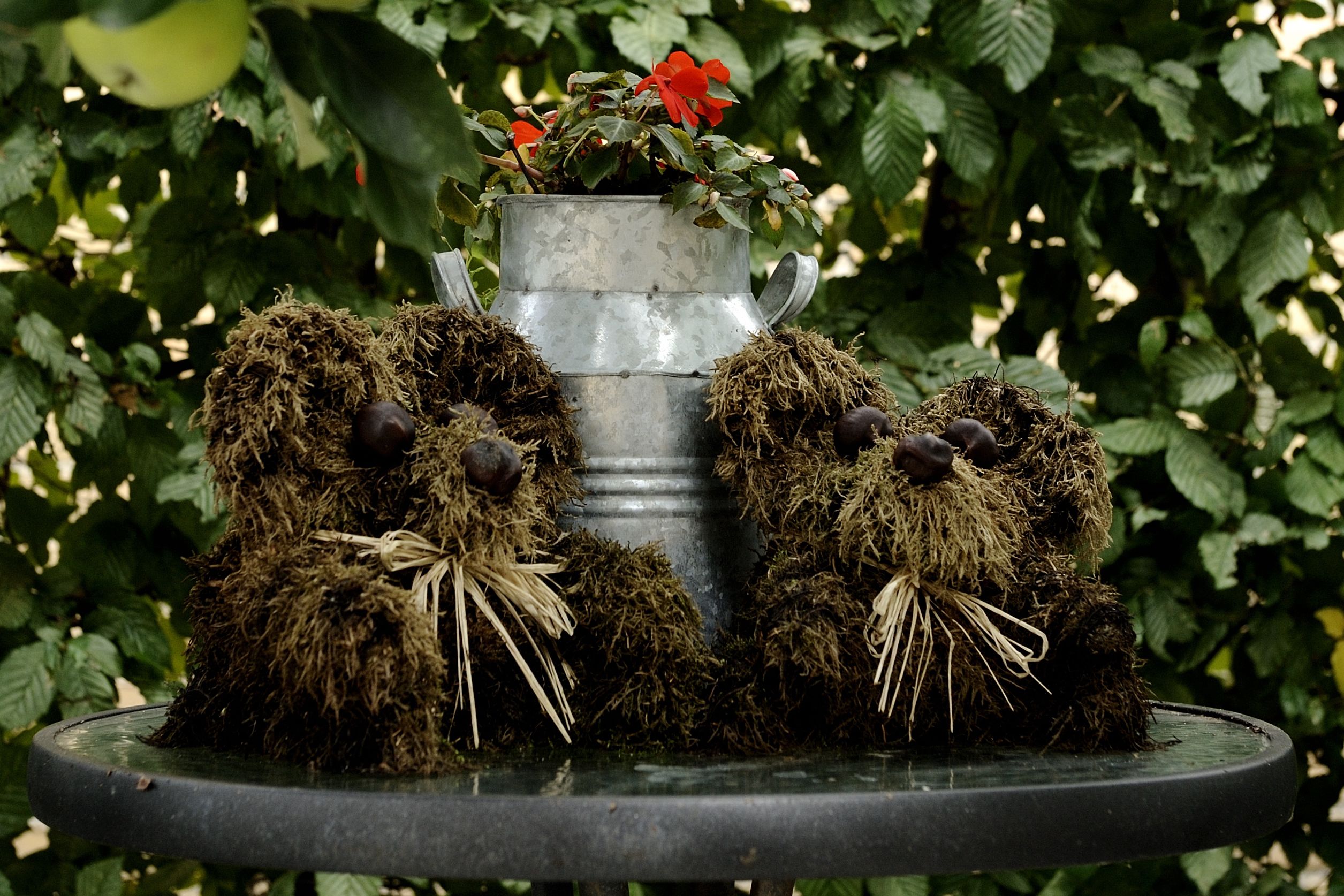 A garden feature with a metal pot containing red flowers, surrounded by moss-covered sculptures.