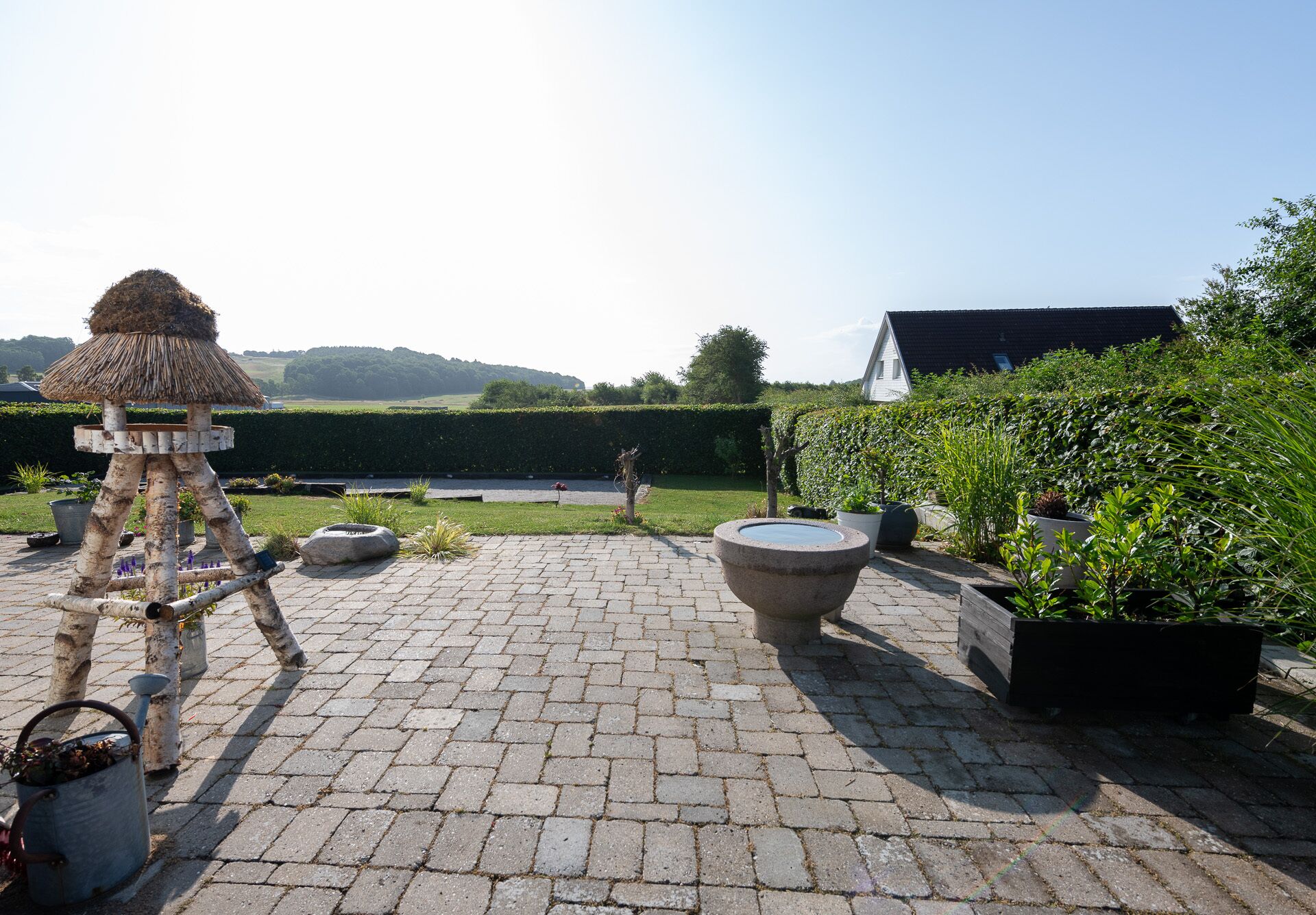 A patio area with a stone floor, a bird feeder, potted plants, and a view of a hedge and distant hills.