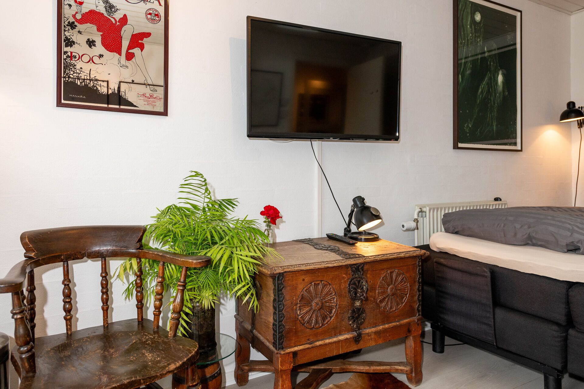 A cozy room with a wooden chair, a small table with a plant, a TV mounted on the wall, and a bed with a grey blanket.
