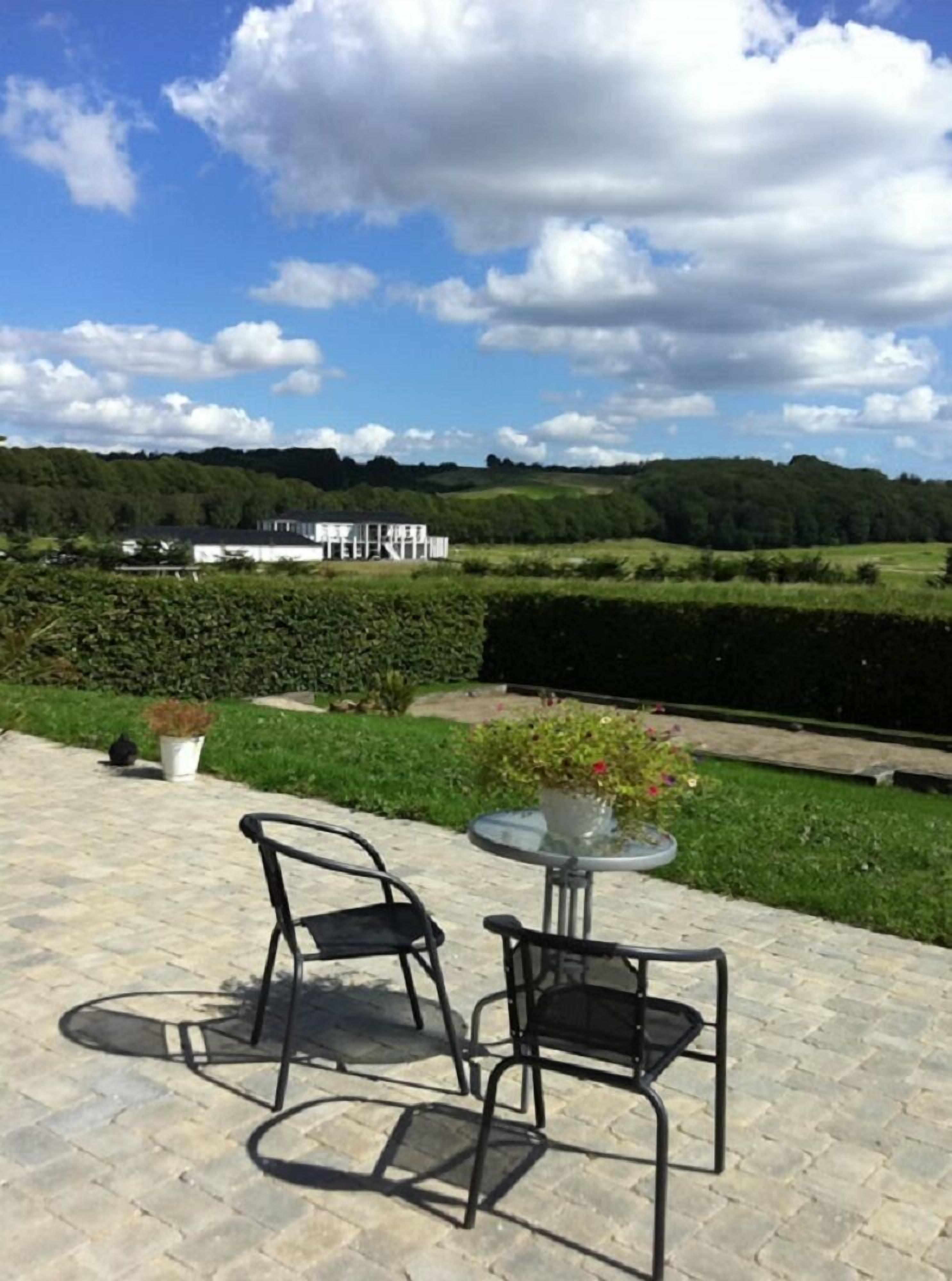 An outdoor patio with two chairs and a table, overlooking a landscaped garden and a white building in the distance.