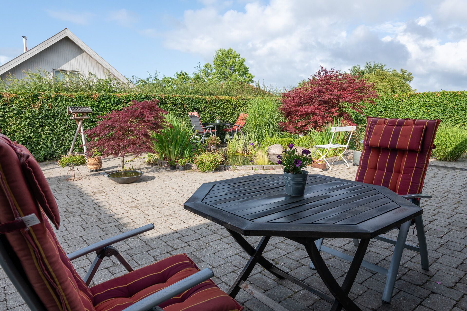 Een terras buiten met een houten tafel en stoelen, omringd door plantenpotten en een kleine vijver.