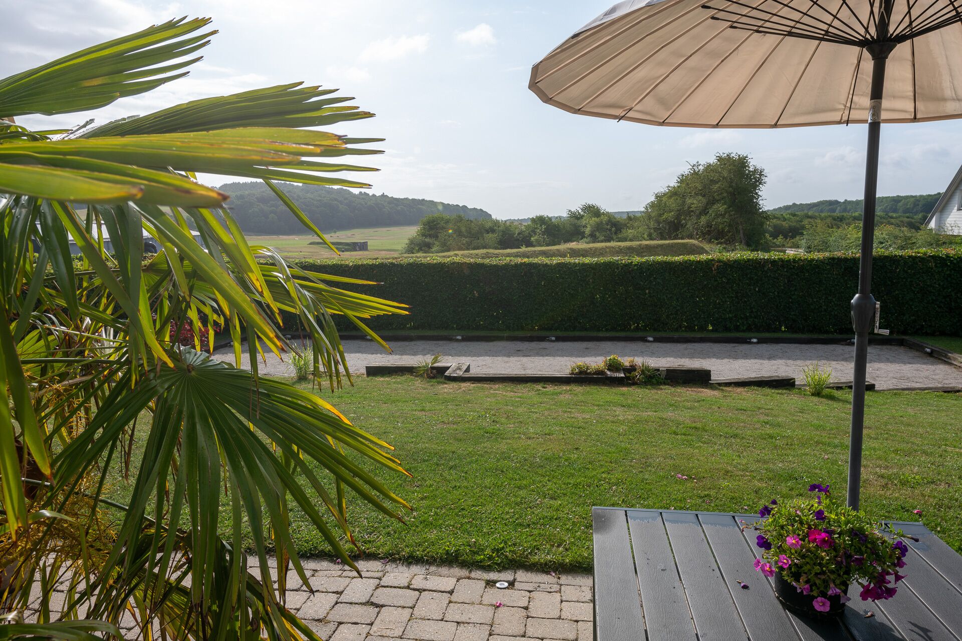 A patio with a wooden table, a beige umbrella, and a flower pot with purple flowers.