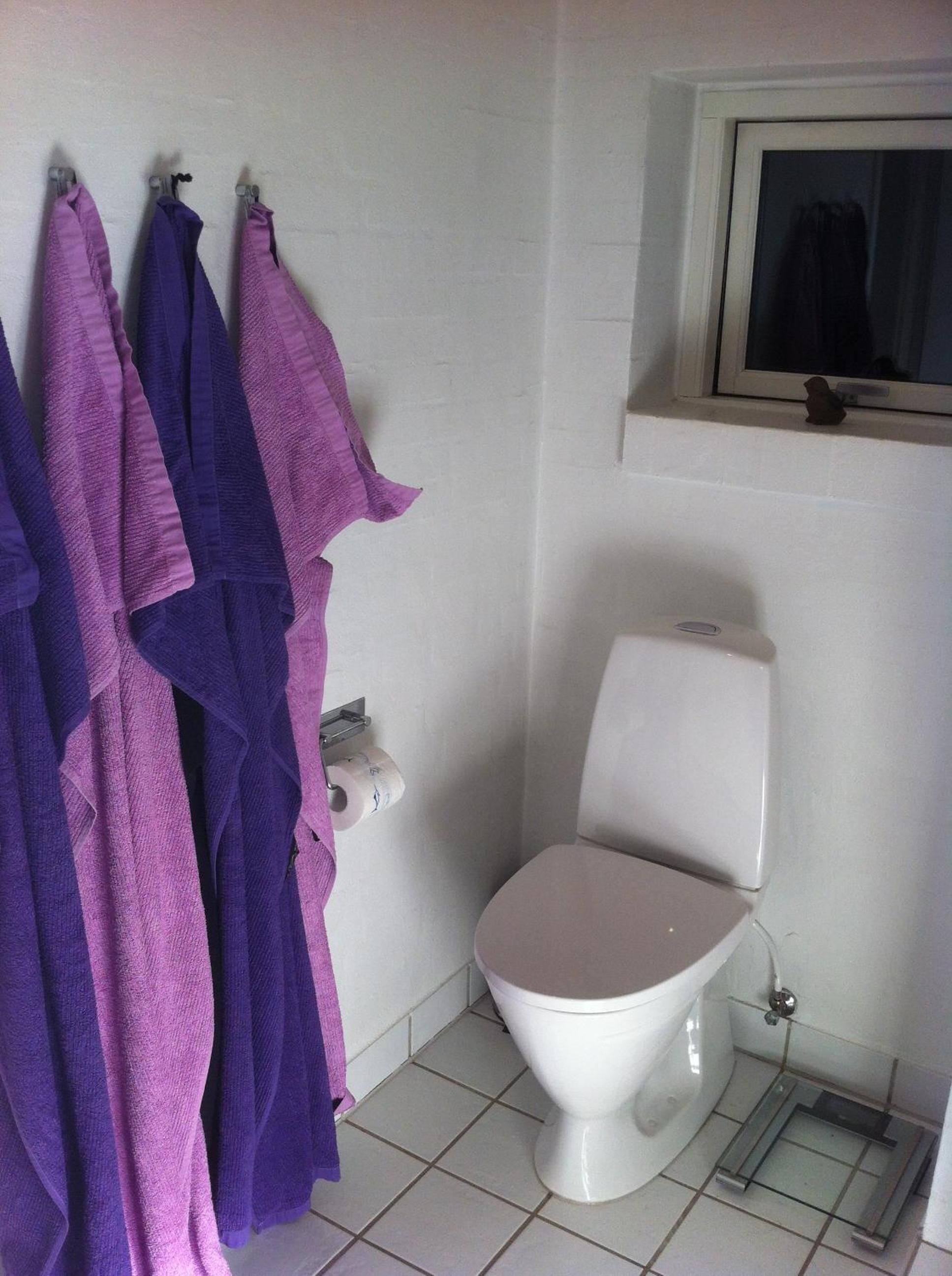 Un baño con un inodoro, una ventana encima y un perchero con toallas moradas.