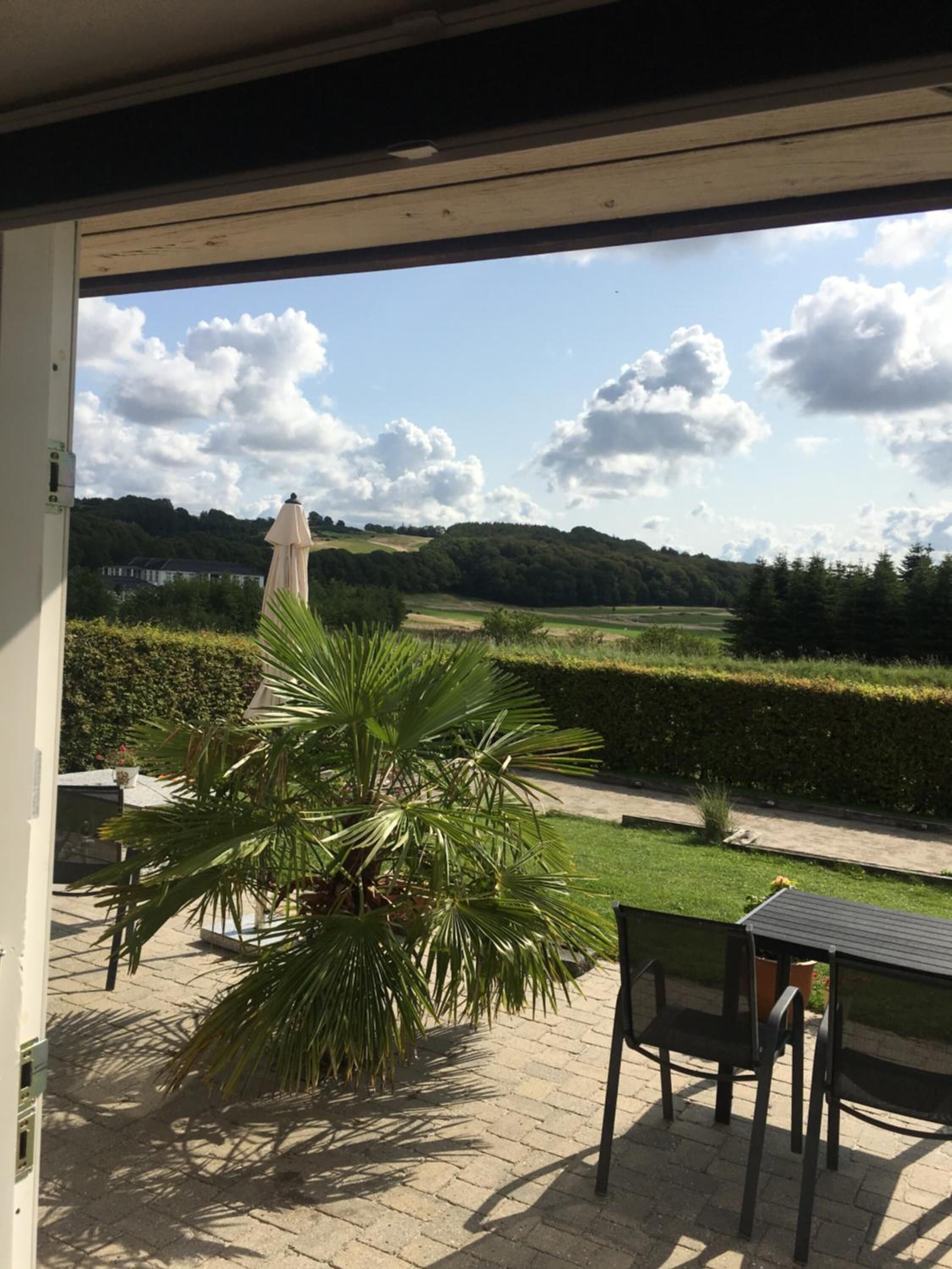 Eine überdachte Terrasse mit Blick auf eine grüne Landschaft, einschließlich einer Hecke und einem Gebäude in der Ferne.