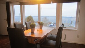 Un comedor con mesa y sillas de madera, ventanas amplias con vista al agua y edificios, y plantas en macetas sobre la mesa.
