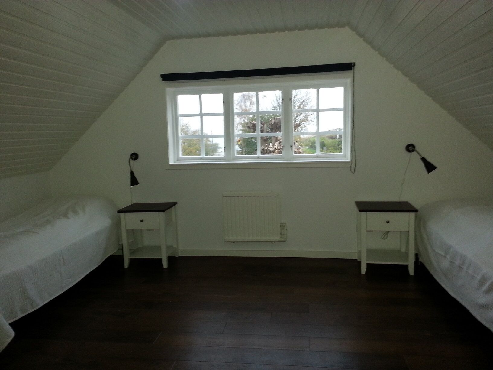 A room with two single beds, a window with white shutters, a radiator, and a small table with a lamp.
