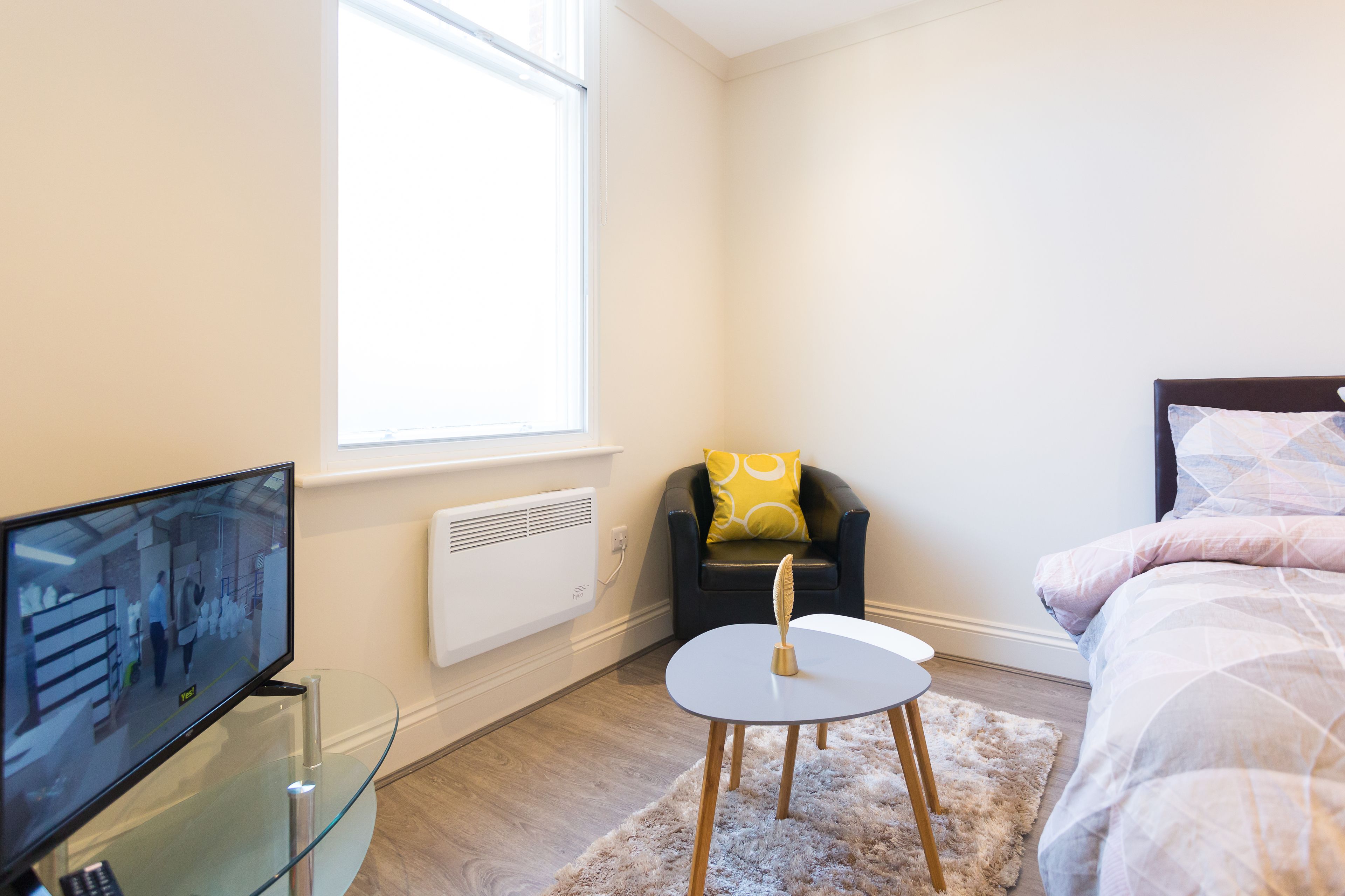 Een moderne slaapkamer met een bed, een stoel, een klein tafeltje en een televisie op een glazen voetstuk.