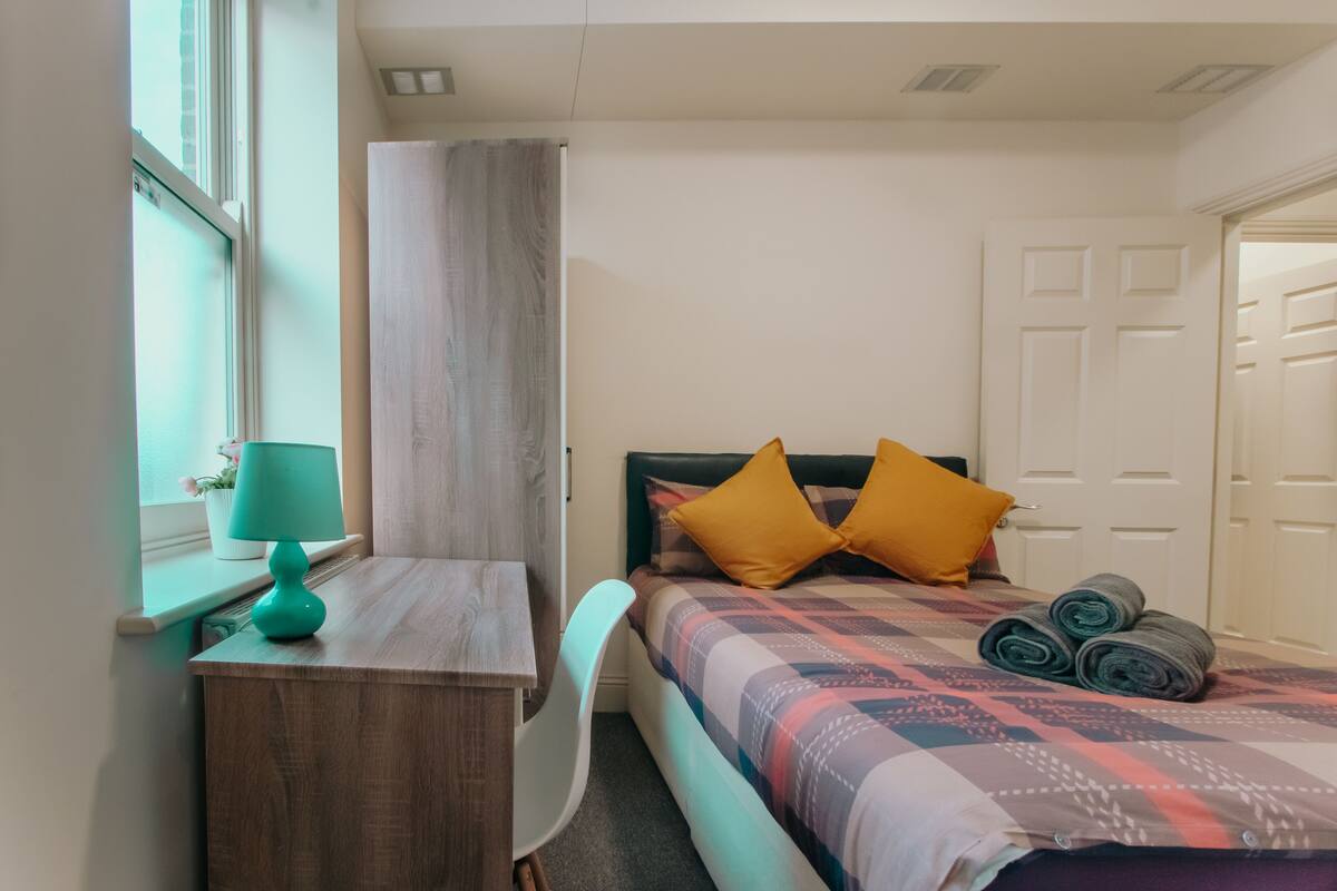 Quarto com cama, escrivaninha, cadeira e janela.