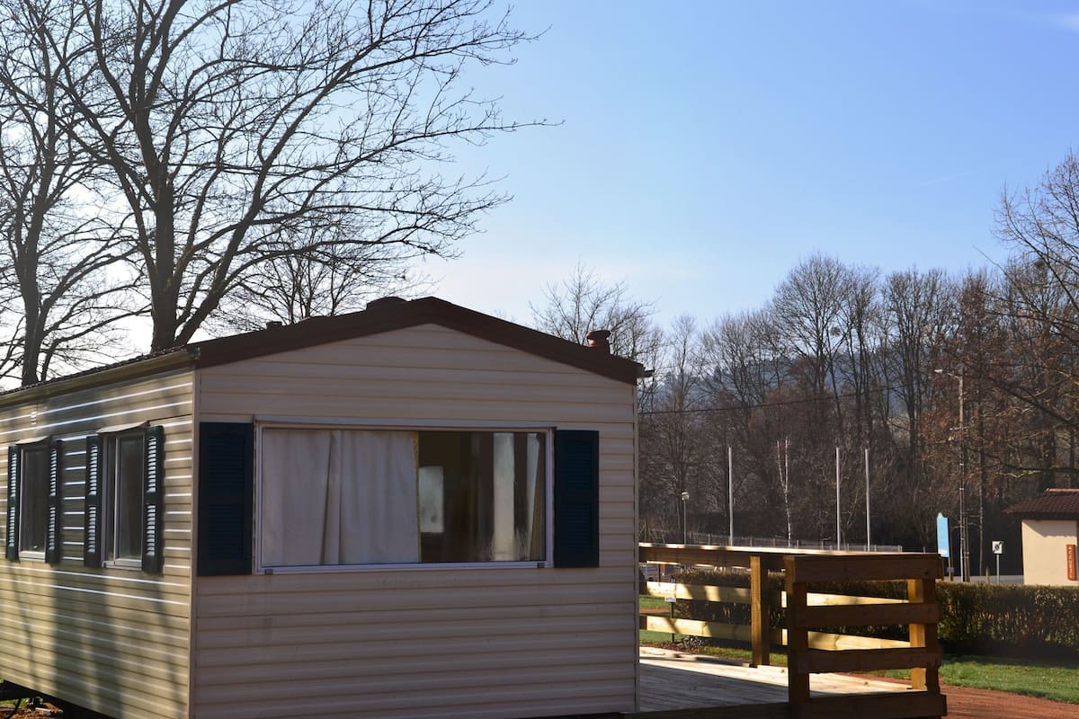 A small, single-story mobile home with a window and shutters, located in a park with a wooden bench and trees in the background.