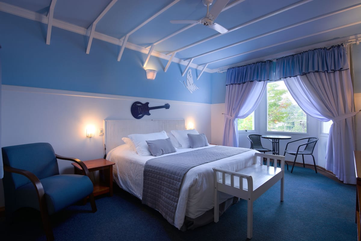 Een slaapkamer met een bed, een stoel, een tafel, een gitaar als wanddecoratie en een plafondventilator.