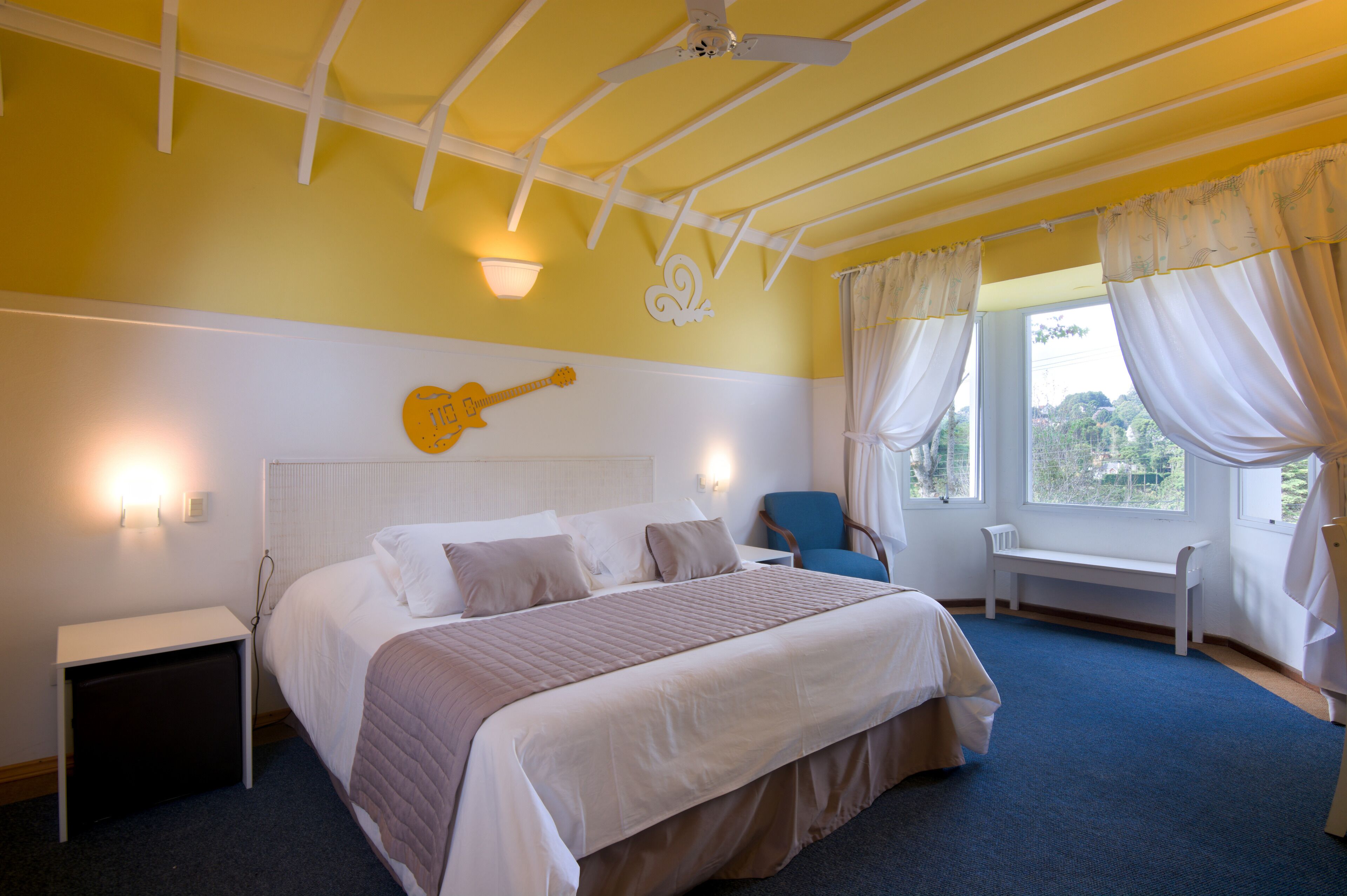 Een slaapkamer met een bed, een gitaar als decoratie aan de muur, een bankje bij het raam en een plafondventilator.