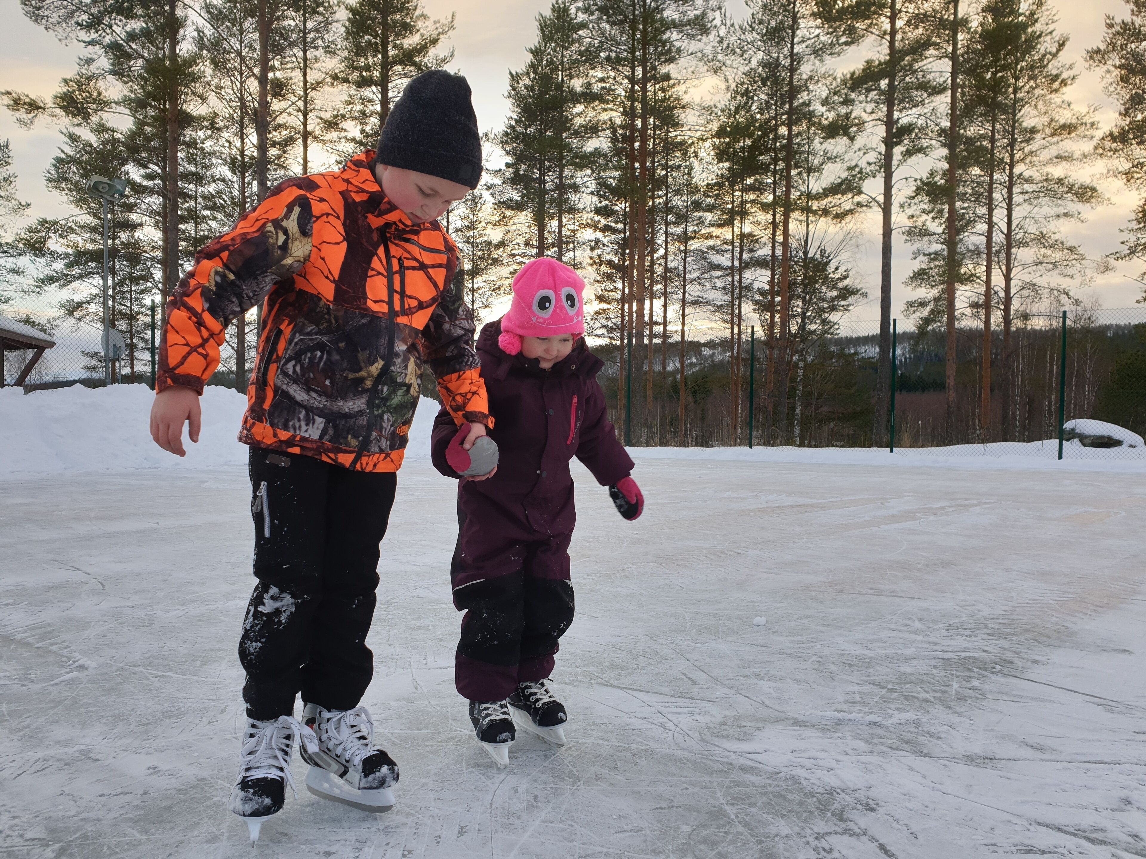 Two children ice skating on a frozen surface with trees in the background.