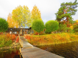 A wooden dock leading to a rustic cabin with stone walls and a chimney, surrounded by autumn foliage and a small pond. - Tahko-Tours (Kuopio)