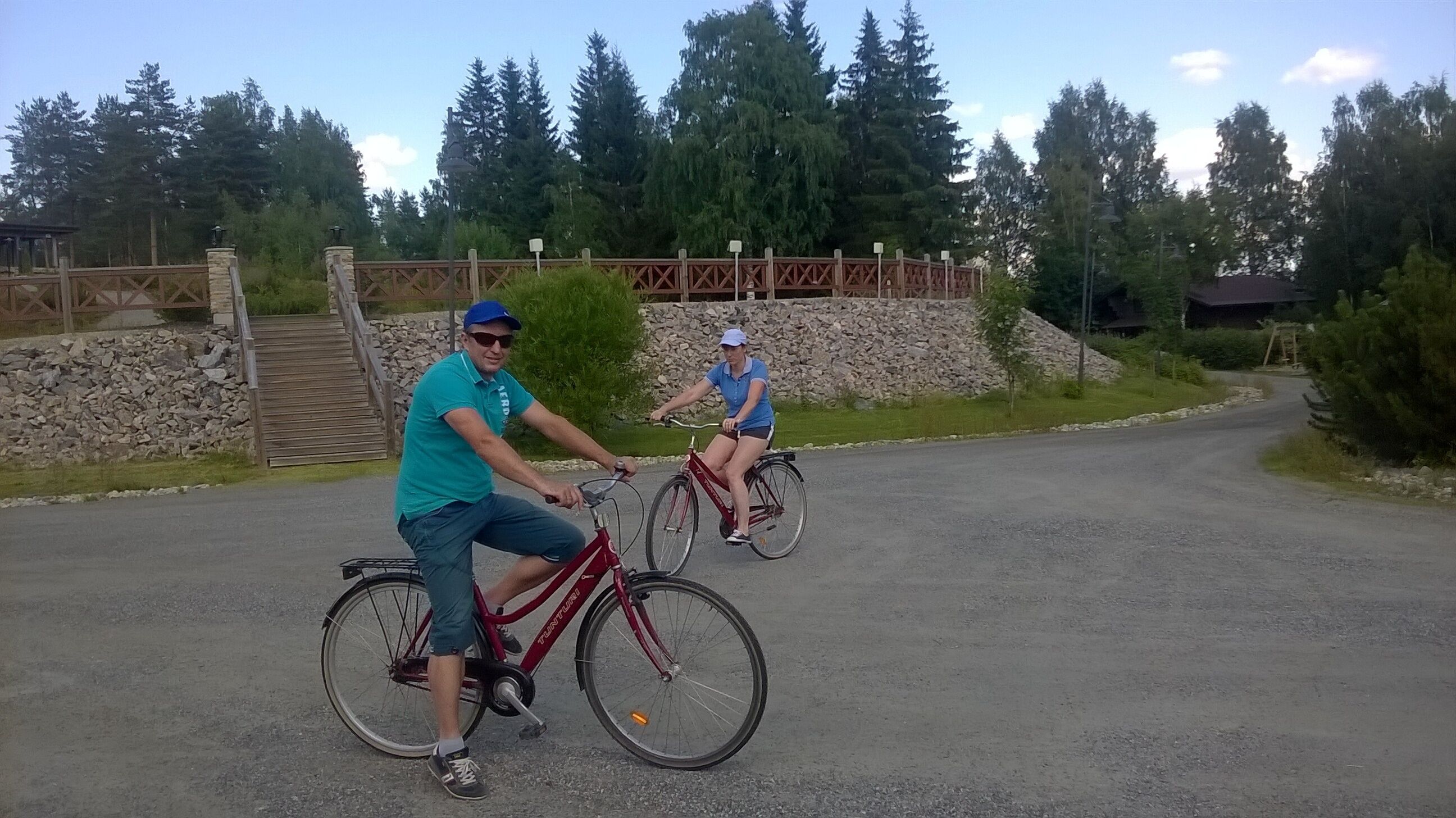 Dos personas montando en bicicleta por una carretera pavimentada, con un muro de piedra y árboles al fondo.
