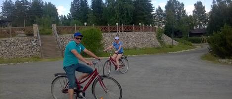Dos personas montando en bicicleta por una carretera pavimentada, con un muro de piedra y árboles al fondo.