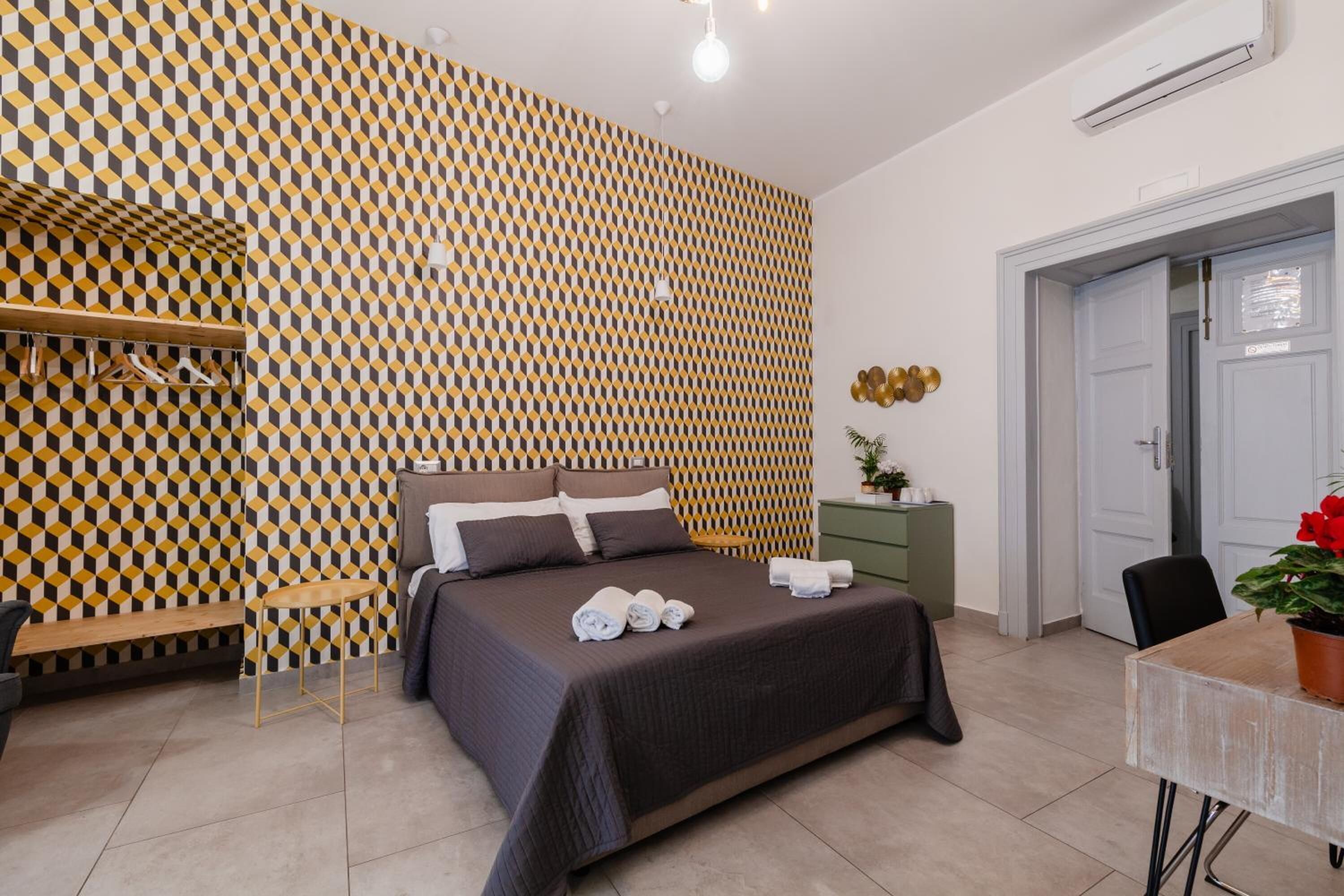 A hotel room with a patterned wall, a bed with towels, a bench, a small table, and a door.