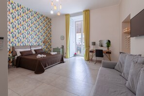 Una habitación de hotel moderna con una pared de diseño geométrico, una cama con ropa de cama marrón, un sofá gris y un pequeño escritorio con una silla.