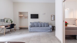 A modern, well-lit room with a grey sofa, a wooden desk with a chair, a small table, and a wall-mounted mirror. - Monteoliveto 33 (Naples)