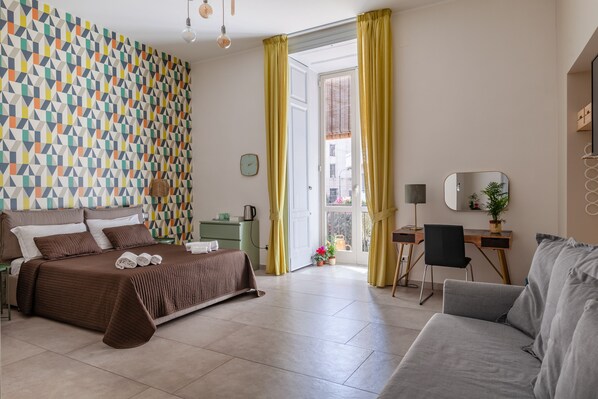 Habitación de hotel con pared estampada, cama con cubierta marrón, sofá gris, escritorio con silla y ventana con cortinas amarillas.