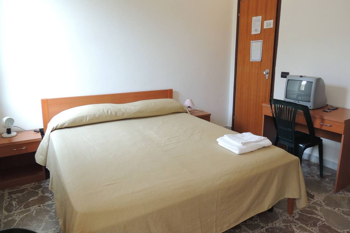 A neatly made bed with a beige bedspread, a wooden nightstand with a lamp, a desk with a computer and chair, and a door with a sign.