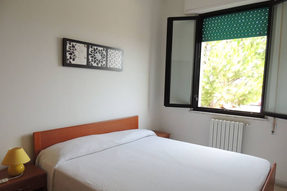 A bedroom with a bed, a nightstand, a lamp, a window with a view of greenery, and a radiator.