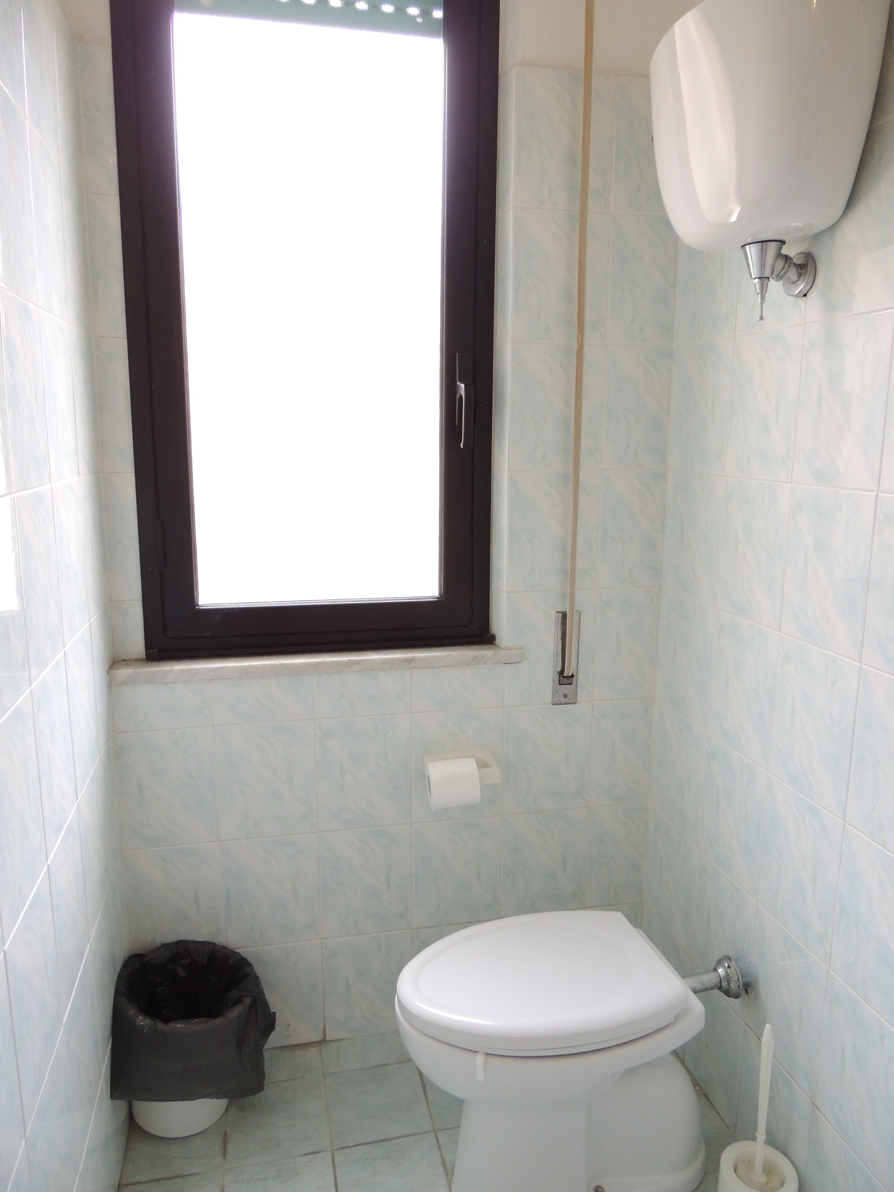Ein kleines Badezimmer mit Toilette, Fenster, Mülleimer und Toilettenpapierhalter.