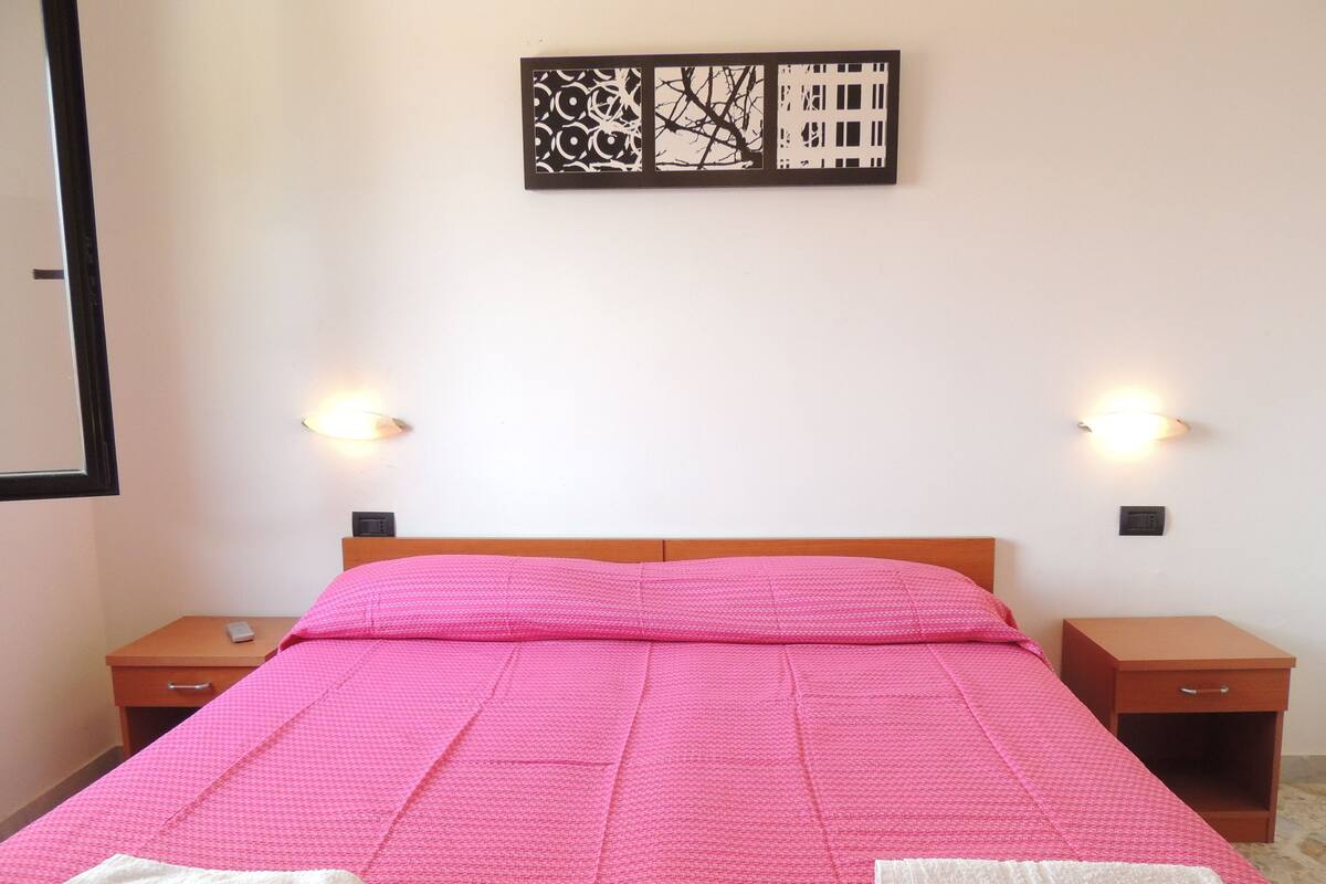 A hotel room with a bed, two bedside tables, a wall-mounted artwork, and two wall-mounted light fixtures.