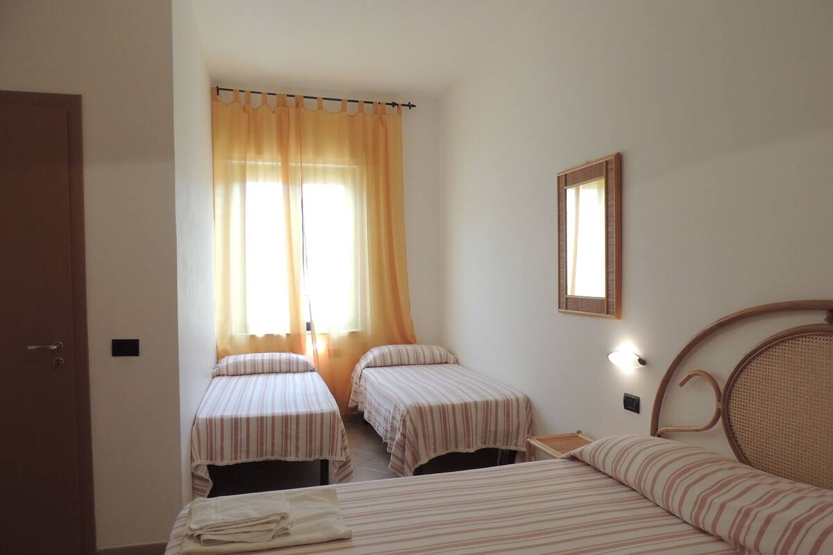 A hotel room with two single beds, a larger bed, a window with curtains, a wall-mounted mirror, and a door.
