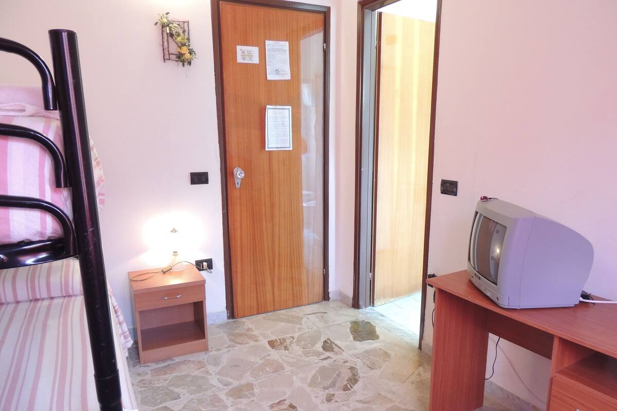 A small room with a wooden door, a small table with a lamp, a television on a wooden stand, and a stone-tiled floor.