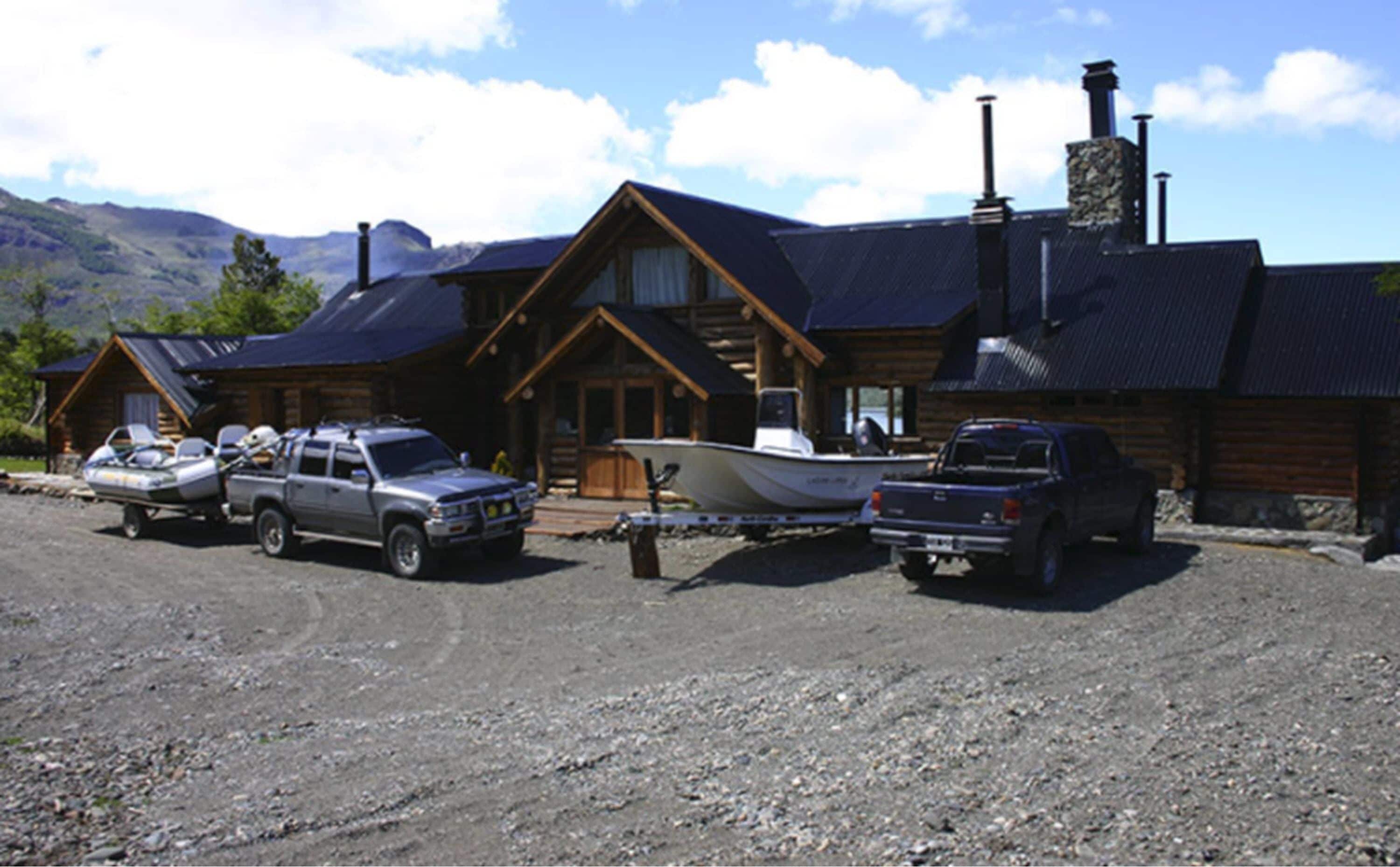 A cabin with a gravel driveway, a boat docked outside, and two trucks parked in front.