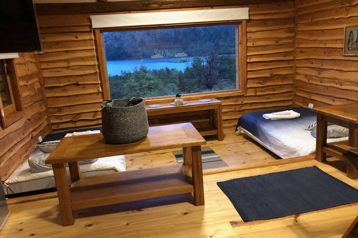 A cozy cabin with a large window offering a scenic view of a forest and lake. Inside, there is a wooden table, a bed with a blue bedspread, and a small table with a basket on it.