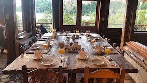 Une table de salle à manger en bois, dressée pour le petit-déjeuner, avec vue sur un paysage boisé grâce à de grandes fenêtres.