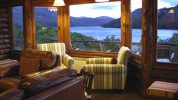 Un intérieur de chalet chaleureux avec un canapé en cuir et des fauteuils à rayures, de grandes fenêtres offrant une vue sur un lac et des montagnes.