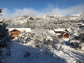 A snow-covered landscape with two wooden cabins, a lake, and a forested hill in the background. - Laguna Larga Lodge (Esquel)