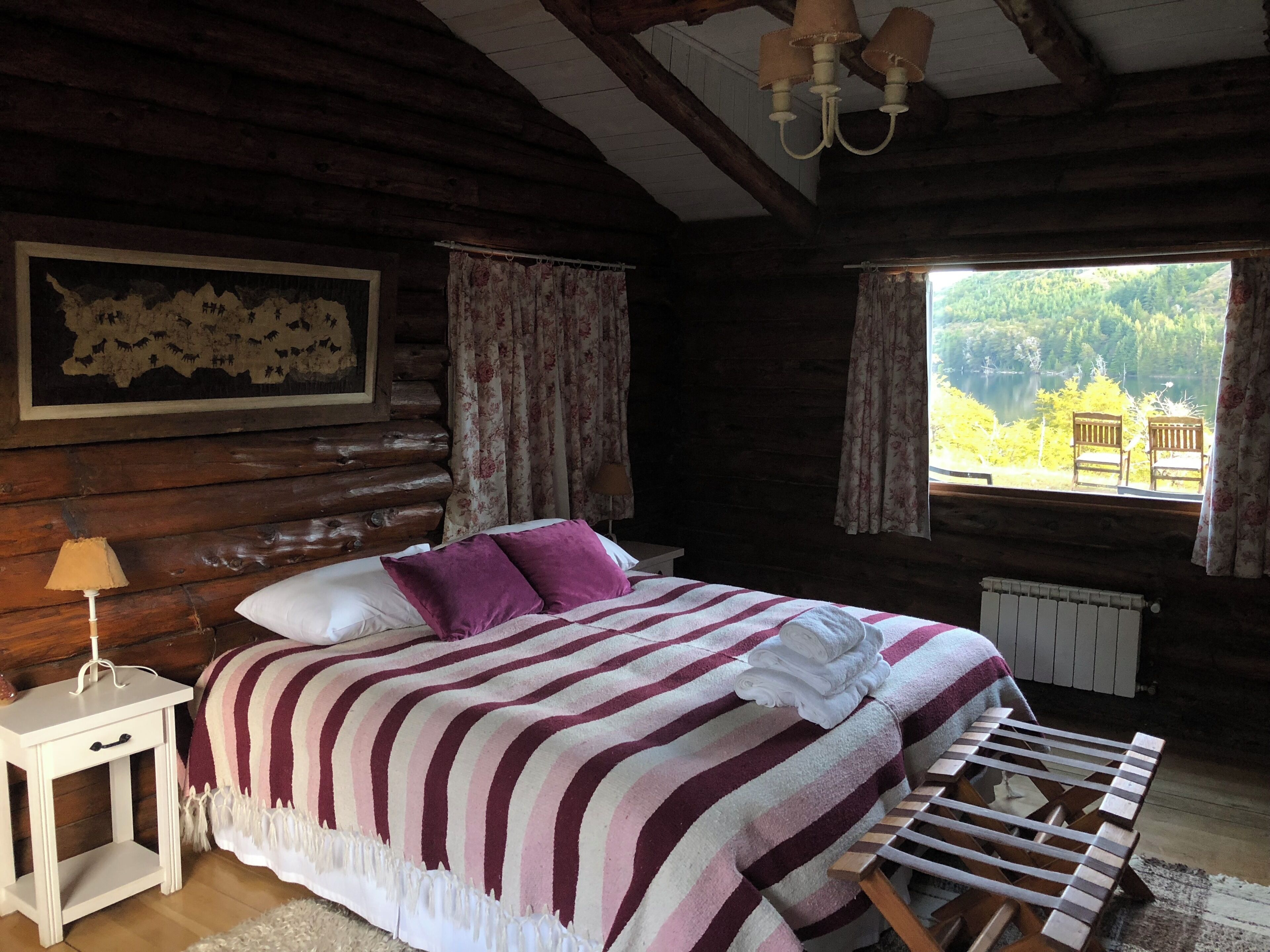 Habitación de cabaña de madera con cama, mesita de noche, ventana con vista a follaje y una silla de madera.