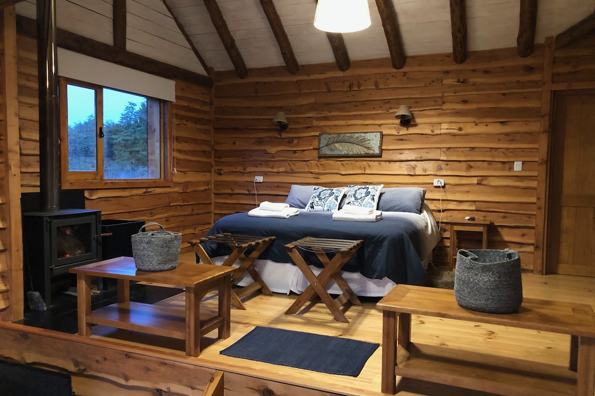 Une pièce chaleureuse aménagée dans un chalet en rondins, avec un lit, une table, un banc, une cheminée et une fenêtre donnant sur la forêt.