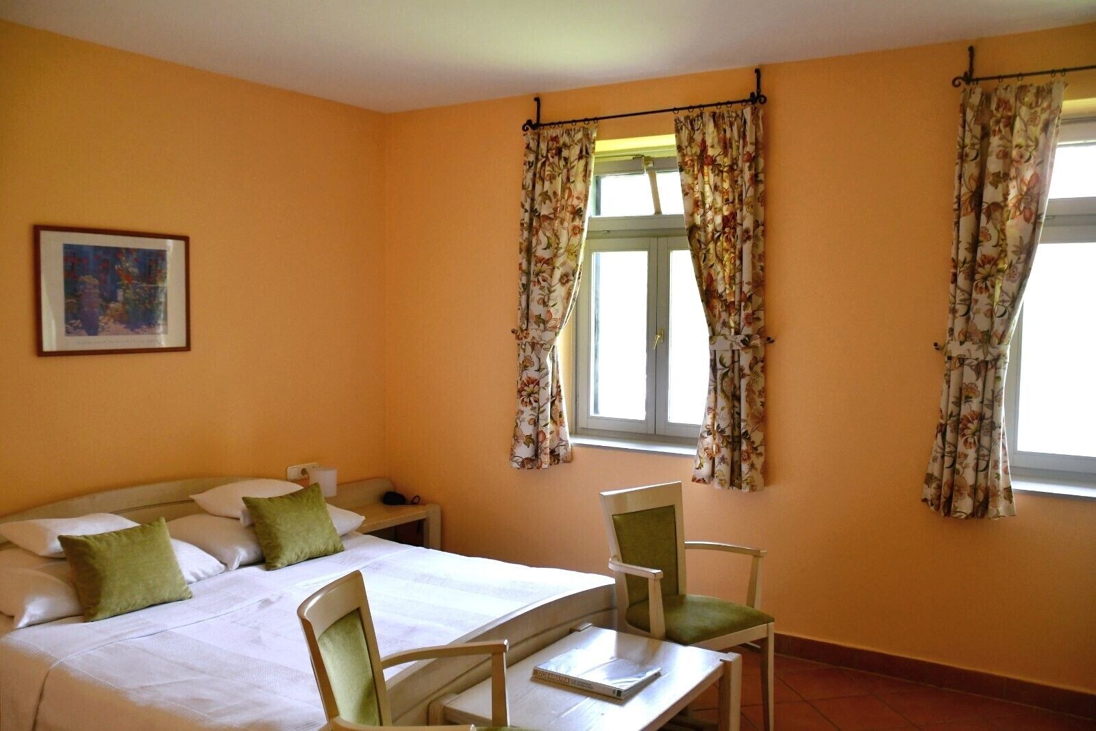 Et hotelværelse med seng, to stole, et lille bord og et vindue med blomstrede gardiner.
