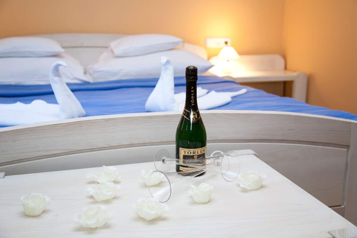 A bed with white pillows and a blue bedspread, a bottle of wine on a table, and a lit bedside lamp.