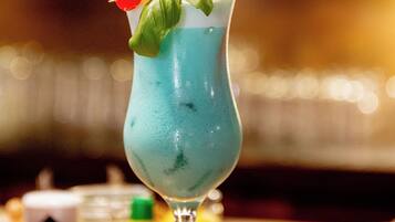 A cocktail in a tall glass with a blue liquid, garnished with a cherry, basil leaves, and a lemon twist, with two straws.